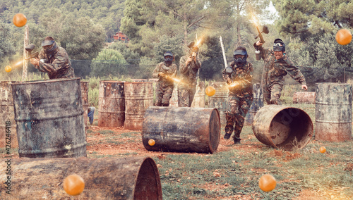 Team of cheerful smiling adult people playing paintball on battlefield outdoor, running with guns
