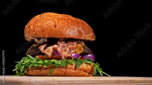 Craft burger is cooking on black background. Consist: sauce, arugula, tomato, onion, bacon, currant sauce, ricotta cheese, air bun and marble meat.