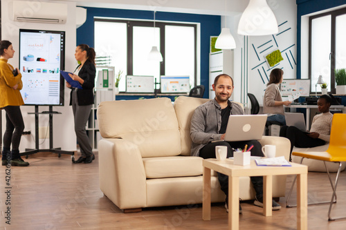 Portrait of man employee typing on laptop looking at camera smiling while diverse team working in background. Multiethnic coworkers talking about startup financial company in modern business office