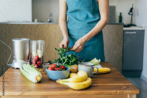 A female cook in a blue apron prepares a vegan fruit cocktail in her home kitchen. Healthy lifestyle and eating concept.