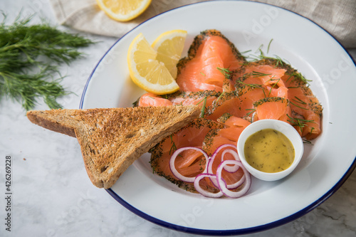 Gravlax Graved Lachs, ein roh gebeizter Fisch mit Dill auf Emaille Teller mit Hovmästarsås Senf Soße, Toast, Zwiebel Ringe, Zitrone und Leinen Tuch auf hell Marmor Hintergrund photo