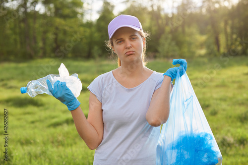 Outdoor portrait of woman volunteer picking up garbage in meadow or park for clean planet, environmentally and ecology friendly, looks at camera with pout lips, holding plastic bottle and garbage bag.