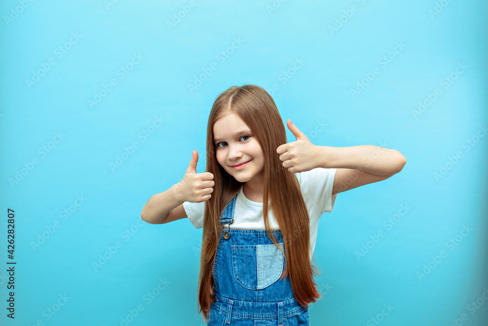Portrait of a smiling girl with a raised thumb on a blue background.