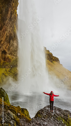 Girl wearing pink jacket standing behind the waterfall with her arms spread wide. She is overjoyed and happy. Huge and powerful waterfall. . Slopes of the mountain overgrown with moss and golden grass