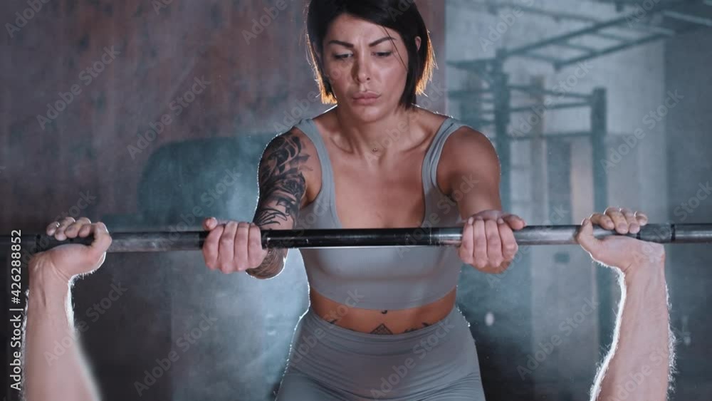 Attractive adult couple training in the gym - athletic man pumping his chest muscles with a dumbbell - a woman hold the dumbbell for the safety Attractive adult couple training in the gym - athletic man pumping his chest muscles with a dumbbell - a woman hold the dumbbell for the safety
