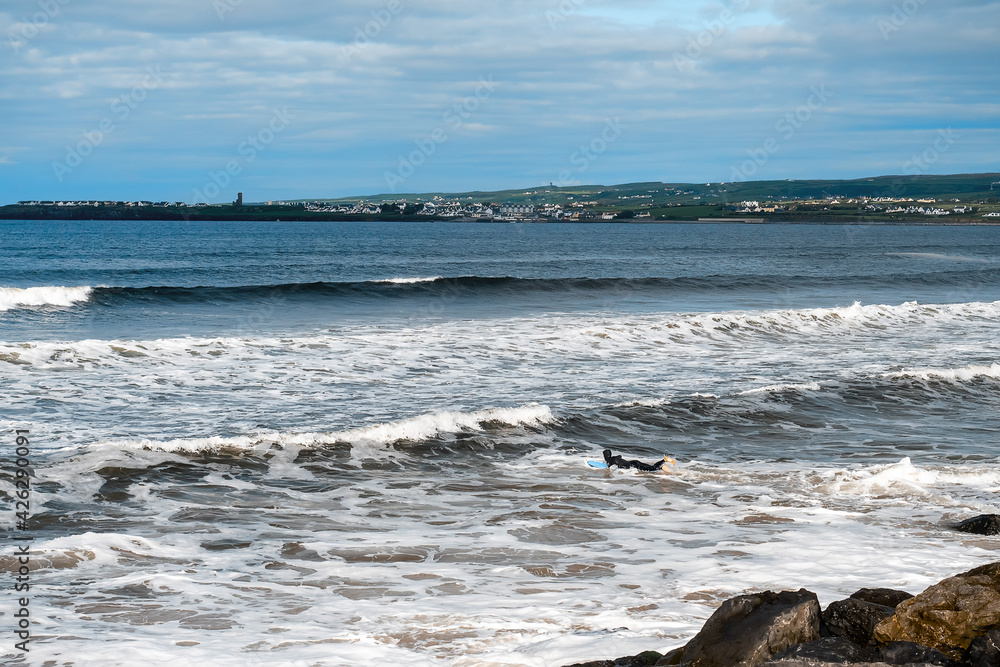 Fototapeta premium Lahinch town coast line. County Clare, Ireland, Sunny day, blue cloudy sky. Nobody. Powerful waves moving towards the beach. Surfer in dark wet suit in the water