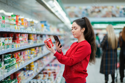 A young woman scans the QR code on a package of yogurt. In the background, a supermarket with visitors in a blur. The concept of modern technologies