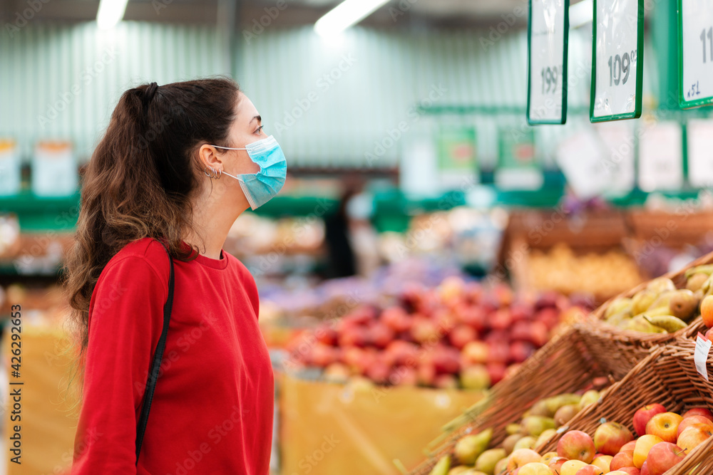 Shopping. Portrait of a young woman in a medical mask on her face looking at apples in a supermarket. The concept of shopping and the new reality