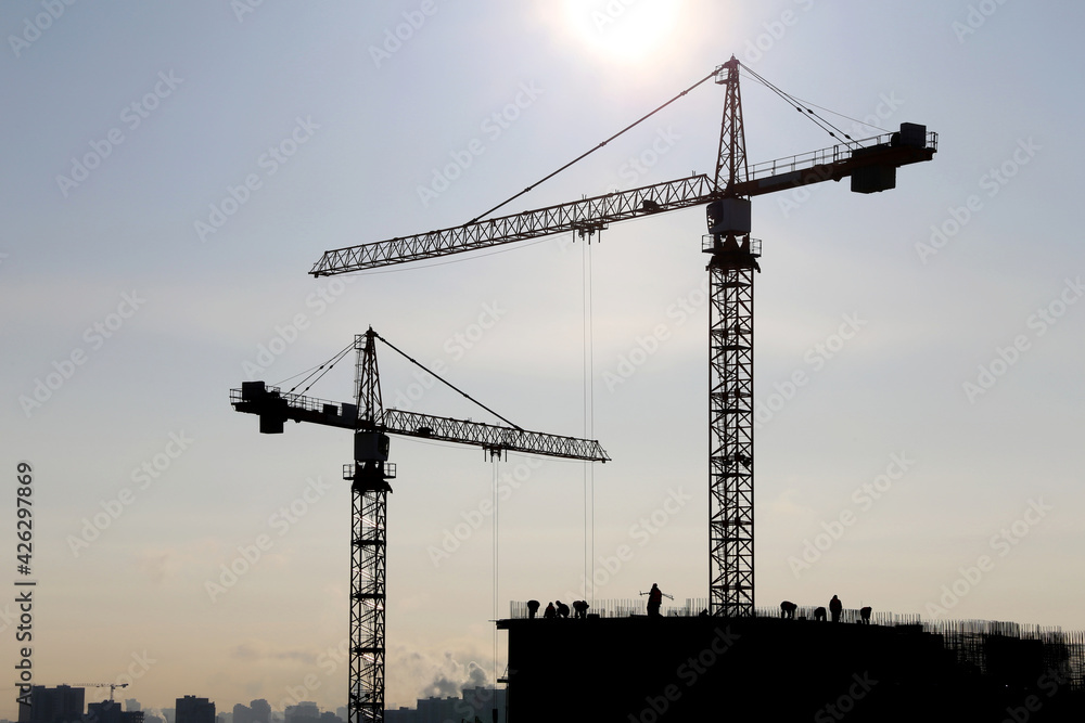 Silhouettes of two tower cranes above the unfinished residential buildings on blue sky and shining sun background. Housing construction, apartment block in city