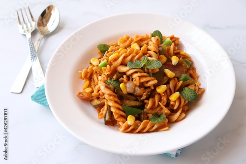 Whole wheat fusilli pasta with vegetables. Vegetable Pasta with zucchini, mushrooms, corn, and peppers on a white plate. Garnished with mint leaves. on a white marble table with cutlery. copy space.