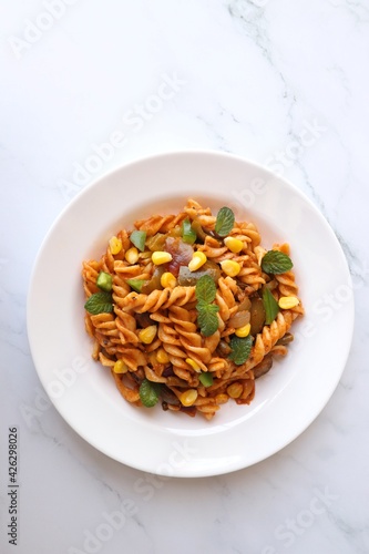 Whole wheat fusilli pasta with vegetables. Vegetable Pasta with zucchini, mushrooms, corn, and peppers on a white plate. Garnished with mint leaves. on a white marble table with cutlery. copy space.