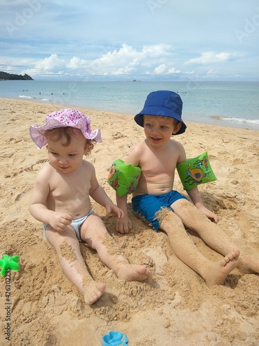 Two preschool children (a boy and a girl) in swimming trunks and panama hats play in the sand on the sea.