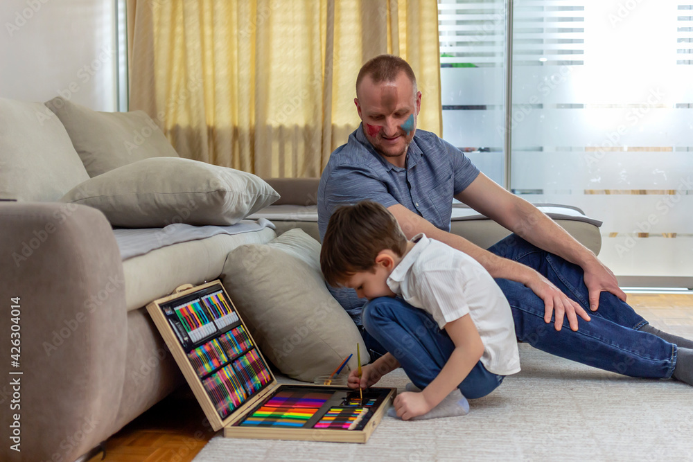 Cute little boy making colorful face on his father. Photo of a young father and his son enjoy the painting at home. Shot of a father and son doing arts and crafts at home. Dad and son having fun.