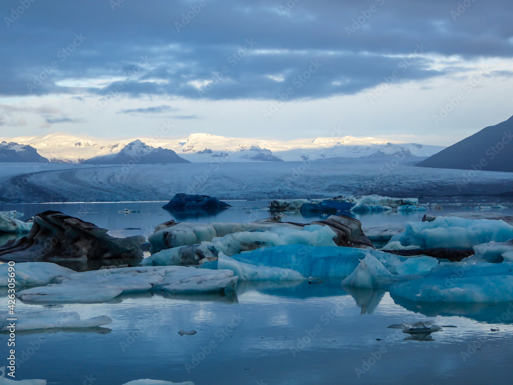 Obraz premium Beautiful glacier lagoon. Thousands of icebergs drifting lazily towards the sea, shining in many shades of blue. Soft sunset in the back. Thick clouds above the lagoon. Glacier's cap in the back