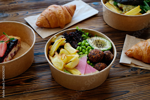 Different takeout food on wooden kitchen table. Hawaiian vegan poke bowl with variety of veggies, french croissant, salmon burger and blueberry cheesecake. Close up, top view, copy space, background.