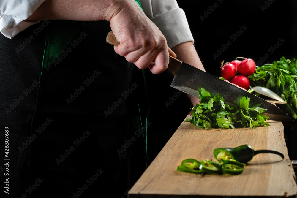 Hands of female chef cutting parsley on wooded board, with radishes on black background. 