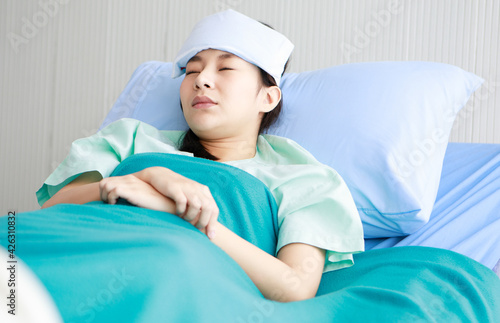 Asian young sick ill girl patient wears green hospital uniform lay down on bed covered by blanket tortured from cold and flu has cool pad gel on forehead to reduce temperature in private wardroom photo