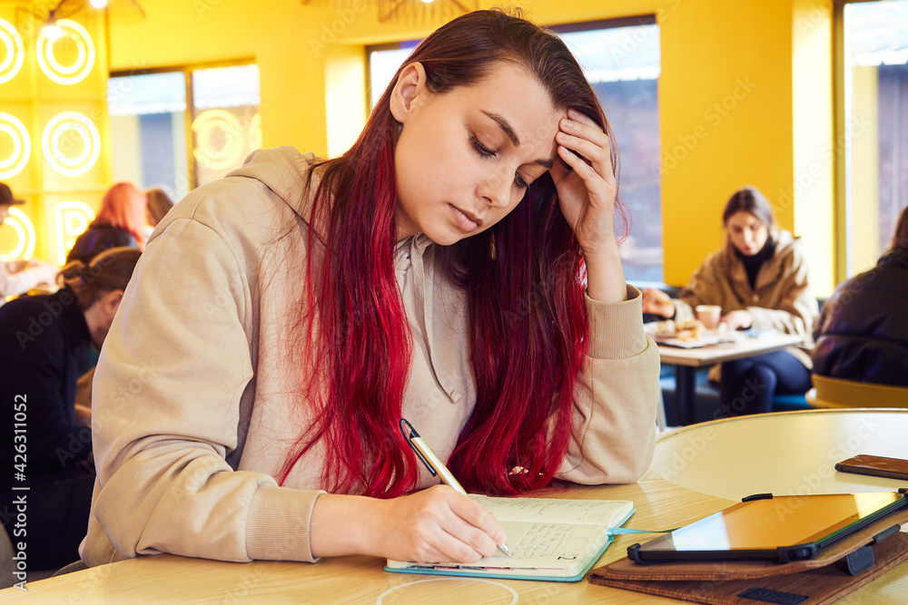 Serious woman with red hair works remotely in a cafe, writes in a diary Serious woman with red hair works remotely in a cafe, writes in a diary