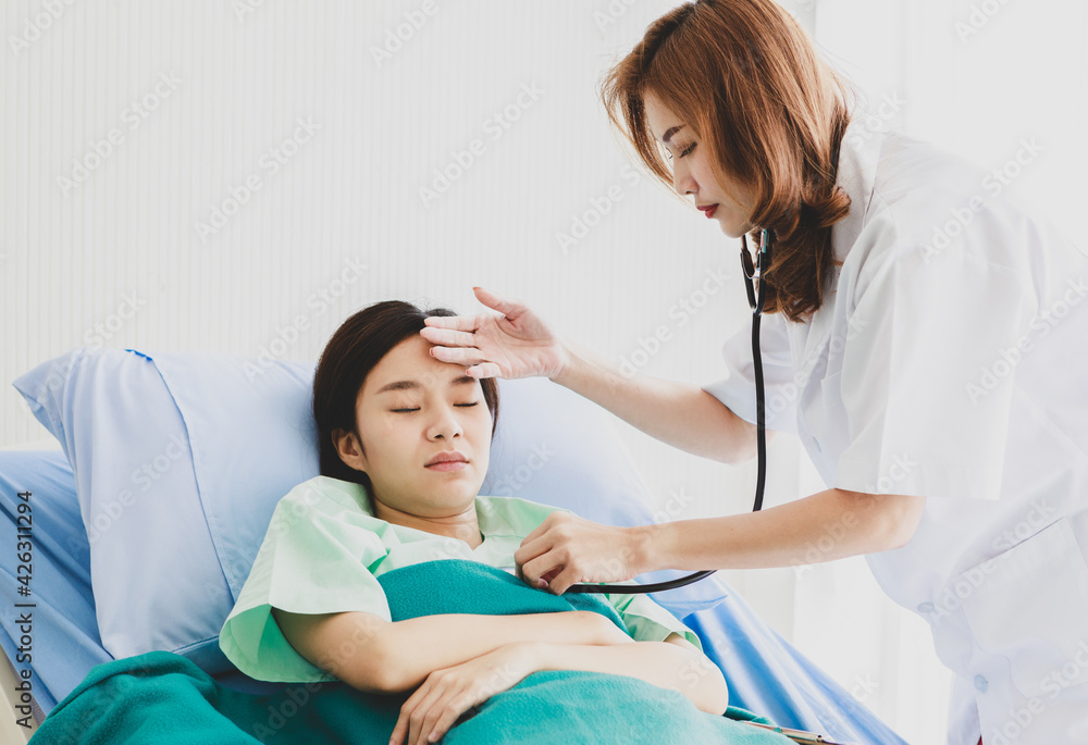 Asian long brown hair female doctor visit patient in green hospital uniform covered by blanket lay down on blue pillow bed check temperature by touching forehead and listen beat pulse by stethoscope Asian long brown hair female doctor visit patient in green hospital uniform covered by blanket lay down on blue pillow bed check temperature by touching forehead and listen beat pulse by stethoscope