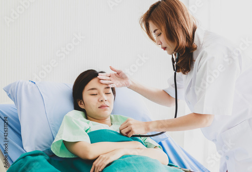 Asian long brown hair female doctor visit patient in green hospital uniform covered by blanket lay down on blue pillow bed check temperature by touching forehead and listen beat pulse by stethoscope