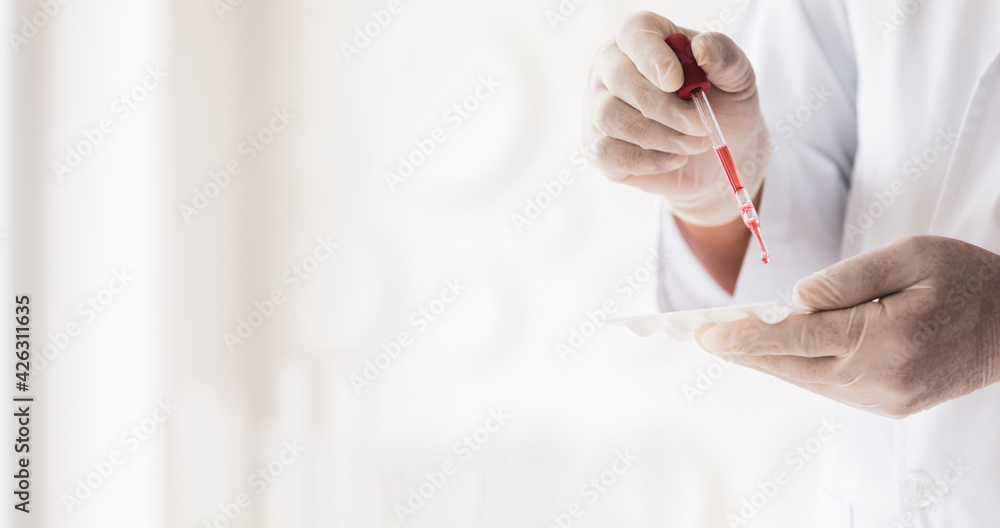 Closeup shot of scientist wears white lab coat and rubber gloves hands holding orange red reagent in glass dropper dropping into plastic plates for experiment in laboratory with blurred background Closeup shot of scientist wears white lab coat and rubber gloves hands holding orange red reagent in glass dropper dropping into plastic plates for experiment in laboratory with blurred background