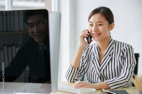 Asian beautiful female employee in black and white stripe shirt sit on chair smiling talk on phone while male worker on opposite office desk who has shadow reflect on computer monitor peek on her photo