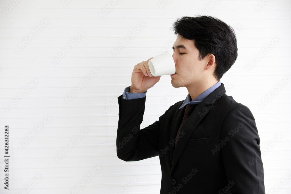 Portrait closeup shot of Asian young successful handsome male businessman wears black formal suit necktie and gray shirt stand look at camera smile hold coffee cup in front white wall background Portrait closeup shot of Asian young successful handsome male businessman wears black formal suit necktie and gray shirt stand look at camera smile hold coffee cup in front white wall background