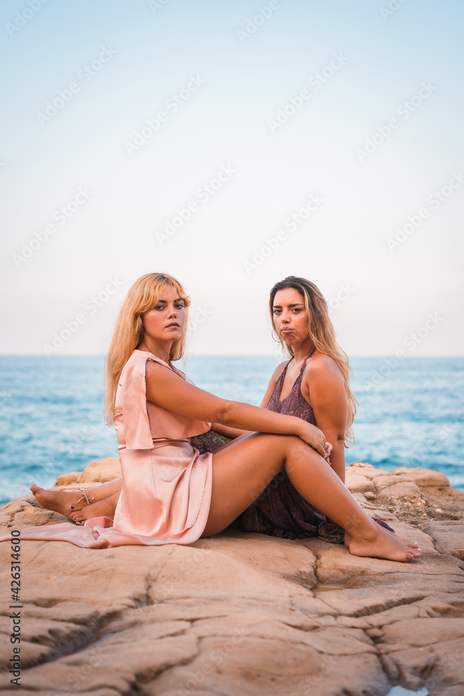 A couple of Caucasian girls sitting in very pretty dresses enjoying the summer by the sea, vertical photo