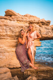 Romantic glances of a couple of Caucasian girls in pink dresses, posing in a summer sunset on the beach on the rocks by the sea