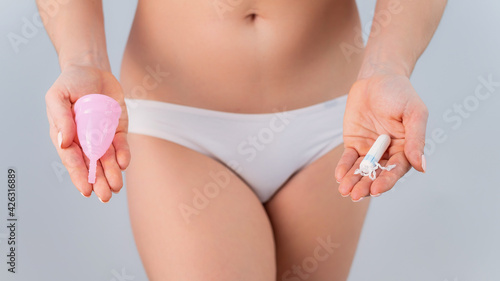 A faceless woman in cotton panties holds a pink menstrual cup and tampon on a white background. Various hygiene products during menstruation