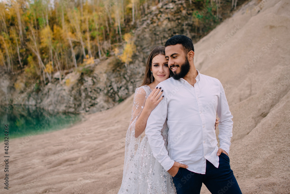 Newlyweds standing near beautiful lake. Middle Eastern groom and Caucasian bride embrace on the beach. Woman presses against her husband's back and looks at camera.
