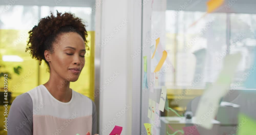 Smiling mixed race businesswoman brainstorming, sticking memo notes on transparent board