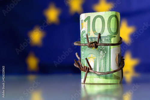 The global economic recession and crisis of European Union. One hundred EURO bancnotes wrapped in barbed wire against flag of EU. Horizontal image.