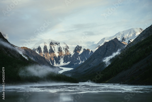 Scenic alpine landscape with snowy mountain peak in golden sunlight and mountain lake in fog under cloudy sky. Atmospheric highland scenery with sunlit mountain top and low clouds on rocks and slopes.