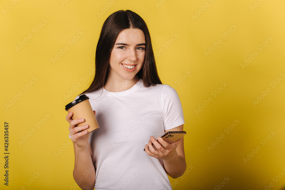 Portrait of attractive girl using phone and hold cup of coffee on yellow background. Place for text. Looking camera. Pretty girl without makeup. Nature beauty Portrait of attractive girl using phone and hold cup of coffee on yellow background. Place for text. Looking camera. Pretty girl without makeup. Nature beauty