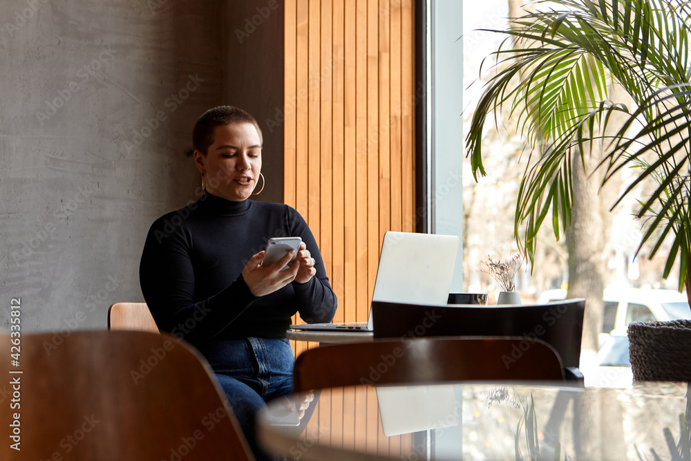 A young girl with short hair and a nose piercing is sitting in a cafe and working on a laptop. The concept of freelancing and remote work or training. A young girl with short hair and a nose piercing is sitting in a cafe and working on a laptop. The concept of freelancing and remote work or training.