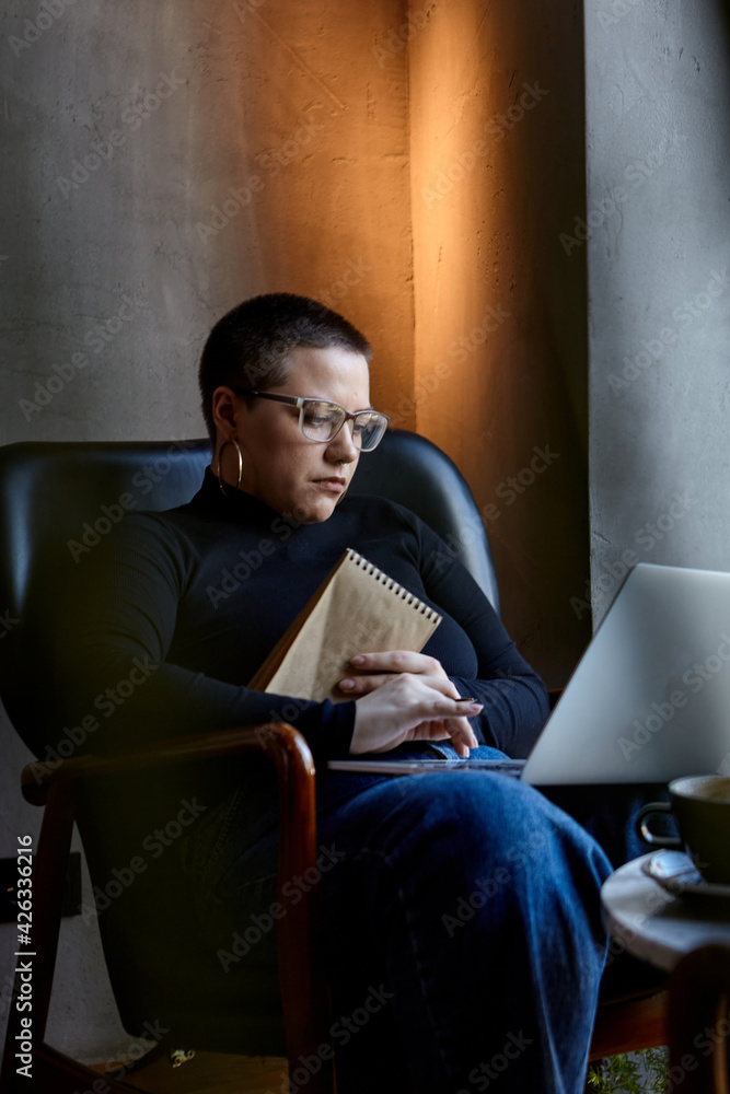 A young girl with glasses, with short hair is sitting in a cafe and working on a laptop. The girl looks at her laptop and makes notes in a notebook. The concept of distance learning A young girl with glasses, with short hair is sitting in a cafe and working on a laptop. The girl looks at her laptop and makes notes in a notebook. The concept of distance learning