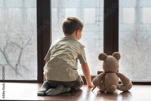 Fotografie Boy and a knitted bear are sitting on the windowsill and looking out the window