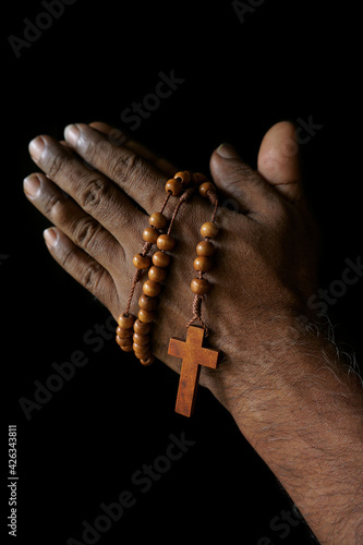 Praying hand of an old Indian Catholic man with wooden rosary isolated on a plain black background. (ID: 426343811)