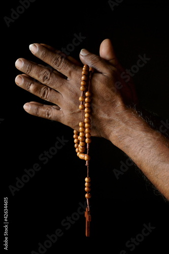 Praying hand of an old Indian Catholic man with wooden rosary isolated on a plain black background. (ID: 426344615)