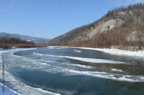 Fototapeta Naklejka Na Ścianę i Meble -  A frozen river in a wintry landscape. Winter landscape with forest, cloudy sky and sun.