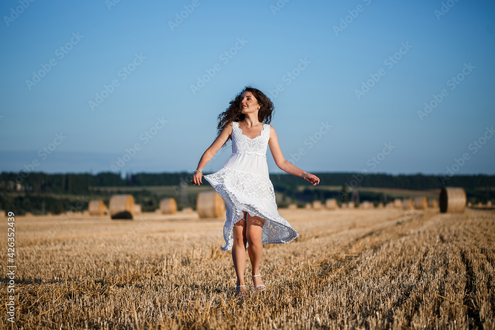 A young beautiful woman in a white summer dress stands on a mown wheat field with huge sheaves of hay, enjoying nature. Nature in the village. Selective focus A young beautiful woman in a white summer dress stands on a mown wheat field with huge sheaves of hay, enjoying nature. Nature in the village. Selective focus