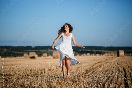Fototapeta Naklejka Na Ścianę i Meble -  A young beautiful woman in a white summer dress stands on a mown wheat field with huge sheaves of hay, enjoying nature. Nature in the village. Selective focus