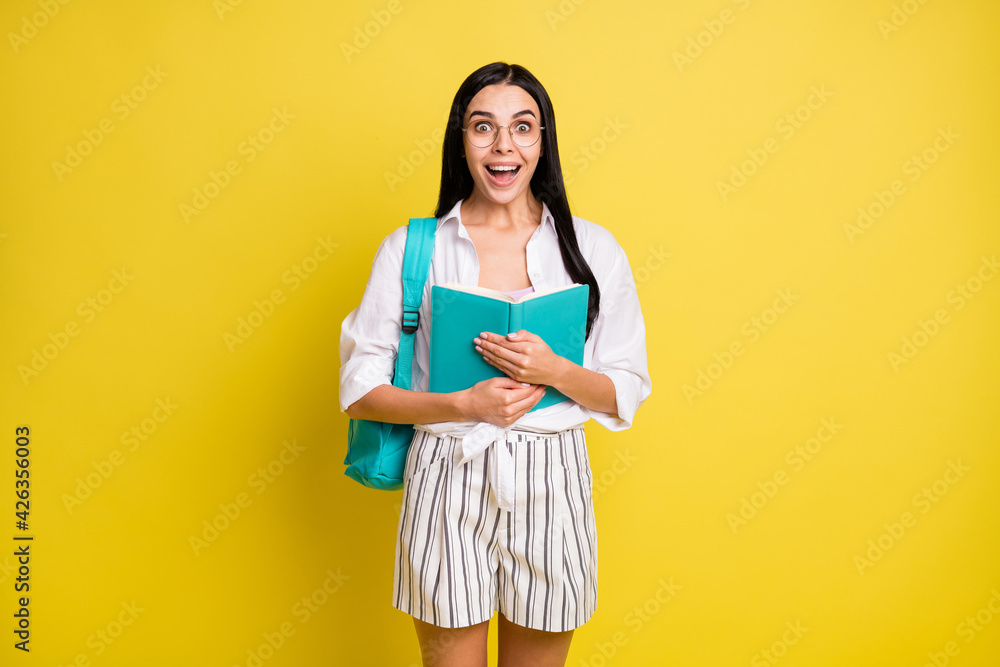 Photo of amazed happy young girl hold book wear backpack glasses isolated on yellow color background Photo of amazed happy young girl hold book wear backpack glasses isolated on yellow color background