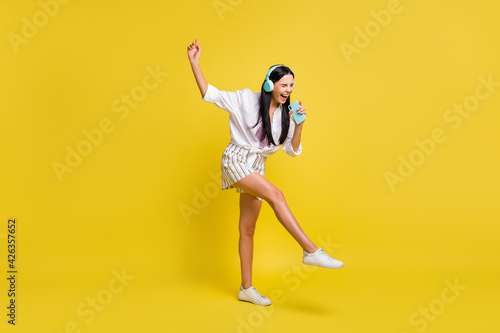 Full size photo of young cheerful smiling positive girl in headphones singing in phone isolated on yellow color background