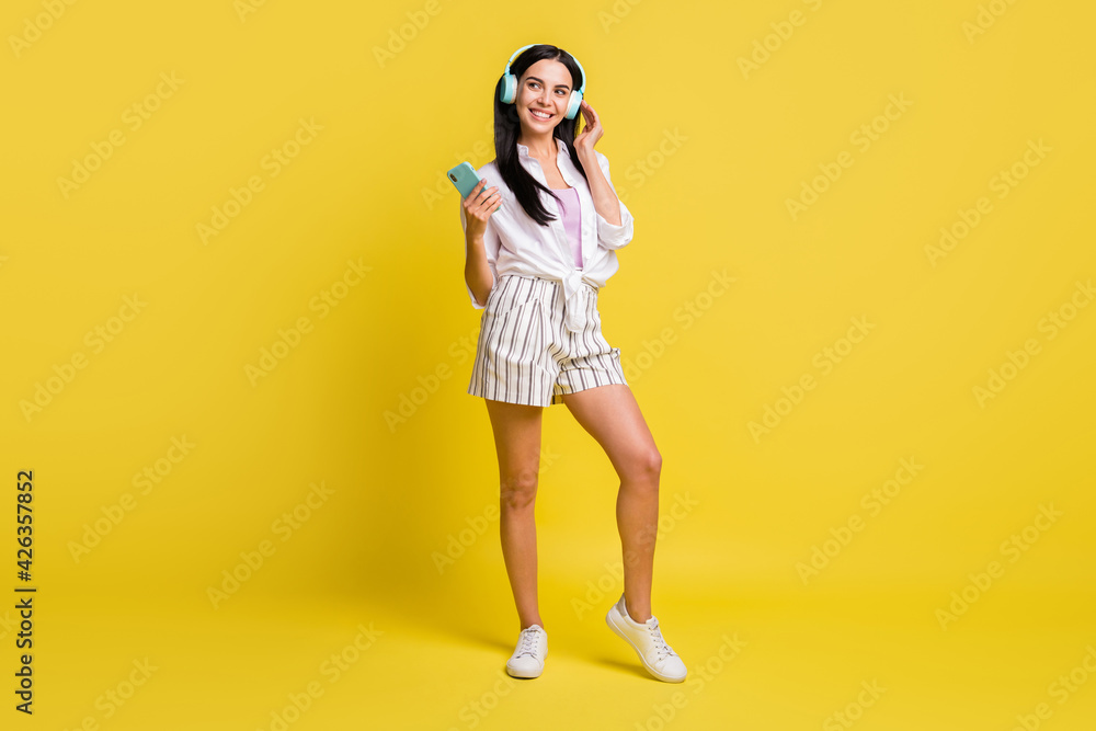 Full length photo of charming positive girl hold phone wear headphones isolated on yellow color background Full length photo of charming positive girl hold phone wear headphones isolated on yellow color background