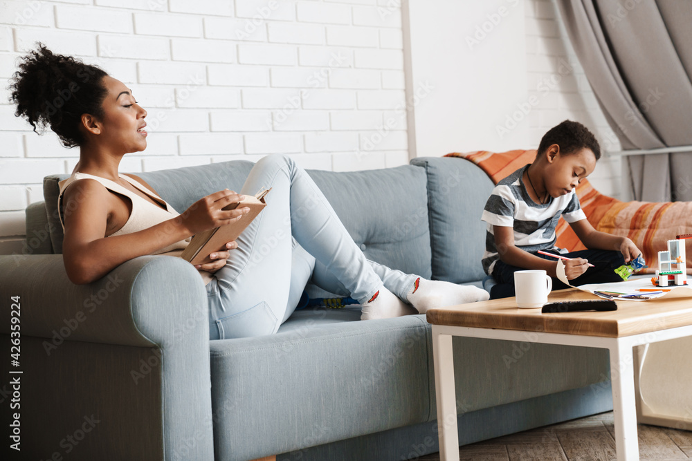 Black woman reading book while her son playing with toys at home Black woman reading book while her son playing with toys at home