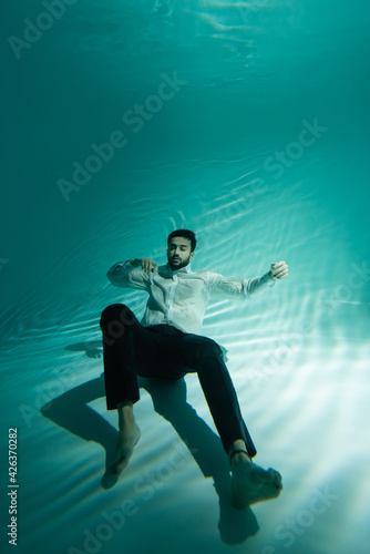 Muslim man in formal wear swimming underwater with closed eyes