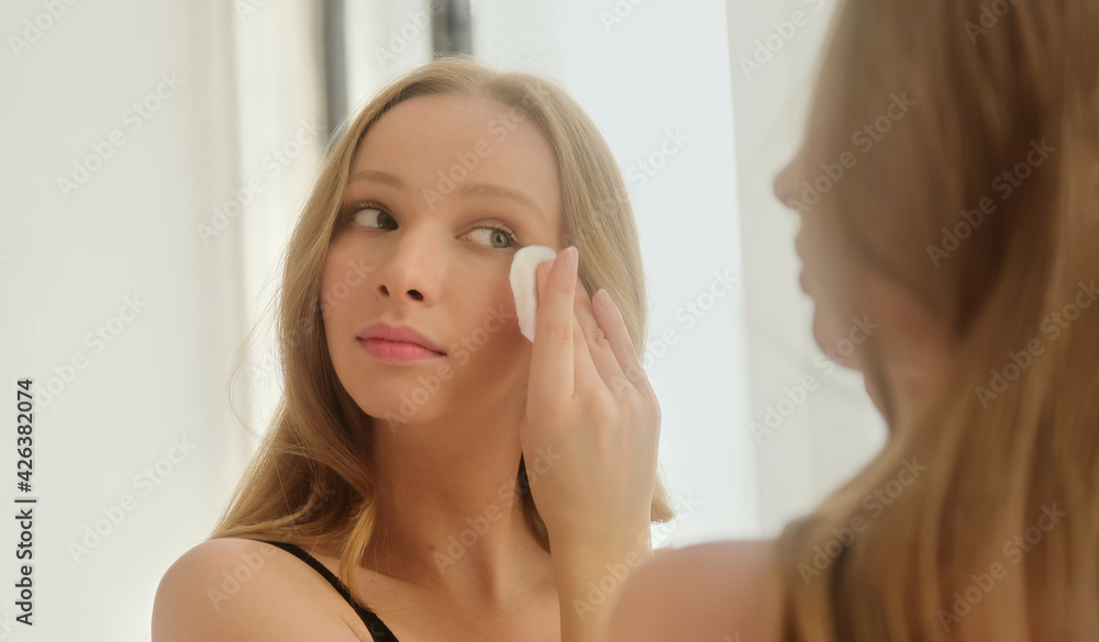 Young woman holding cotton pad disk cleaning face skin with cleanser tonic. Skin care remove make up purify lotion in front mirror at home. Young woman holding cotton pad disk cleaning face skin with cleanser tonic. Skin care remove make up purify lotion in front mirror at home.