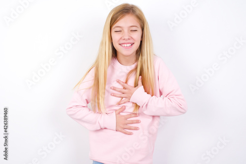 beautiful caucasian little girl wearing pink hoodie over white background keeps hands crossed, laughs out loud at good joke, wears casual clothes.